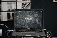 A MacBook Pro laptop displays a textured stone-like wallpaper with Japanese text and the phrase 'Grace Upon Grace' beneath it. The laptop is flanked by two small speakers or green-accented devices. In the background, a dark wall is adorned with various images or posters, mainly architectural in nature.