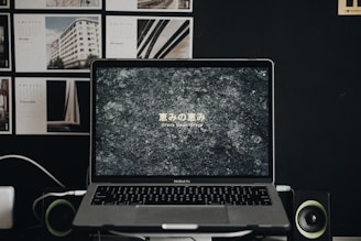 A MacBook Pro laptop displays a textured stone-like wallpaper with Japanese text and the phrase 'Grace Upon Grace' beneath it. The laptop is flanked by two small speakers or green-accented devices. In the background, a dark wall is adorned with various images or posters, mainly architectural in nature.