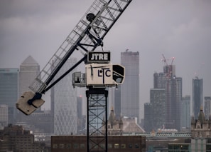 A large crane is positioned in front of a city skyline with tall skyscrapers under a cloudy sky. Many of the buildings have distinctive modern architecture, and there is a mix of construction and finished structures visible.