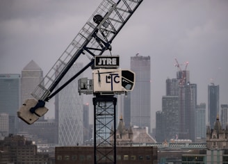 A large crane is positioned in front of a city skyline with tall skyscrapers under a cloudy sky. Many of the buildings have distinctive modern architecture, and there is a mix of construction and finished structures visible.