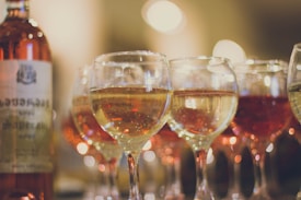 A group of wine glasses filled with various shades of wine is arranged in a row. A wine bottle is positioned on the left side, slightly blurred, with a label containing text. The background is softly out of focus, featuring warm, twinkling lights that contribute to a cozy and inviting atmosphere.