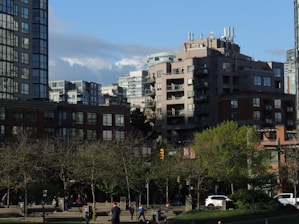 A cityscape featuring modern apartment buildings with large windows and balconies against a backdrop of partly cloudy blue sky. Trees with early spring foliage are visible in the foreground, and people are walking on the pavement, including a person wearing a red hat. A few cars are parked or passing by on the street.