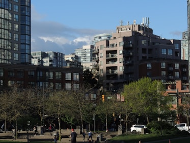 A cityscape featuring modern apartment buildings with large windows and balconies against a backdrop of partly cloudy blue sky. Trees with early spring foliage are visible in the foreground, and people are walking on the pavement, including a person wearing a red hat. A few cars are parked or passing by on the street.