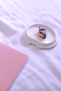 A single piece of chocolate-covered candy with a pink filling sits on a white ceramic saucer on a soft, white fabric surface. In the bottom left corner, there is a pink notebook partially visible.