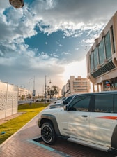 A parked white SUV with the model designation 4800 VTC is in the foreground, positioned on a paved walkway beside a grassy area. The scene is urban, featuring modern buildings and streetlights with decorative design. The sky is partly cloudy with the sun casting warm light, suggesting either morning or late afternoon.
