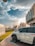 A parked white SUV with the model designation 4800 VTC is in the foreground, positioned on a paved walkway beside a grassy area. The scene is urban, featuring modern buildings and streetlights with decorative design. The sky is partly cloudy with the sun casting warm light, suggesting either morning or late afternoon.