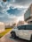 A parked white SUV with the model designation 4800 VTC is in the foreground, positioned on a paved walkway beside a grassy area. The scene is urban, featuring modern buildings and streetlights with decorative design. The sky is partly cloudy with the sun casting warm light, suggesting either morning or late afternoon.