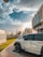 A parked white SUV with the model designation 4800 VTC is in the foreground, positioned on a paved walkway beside a grassy area. The scene is urban, featuring modern buildings and streetlights with decorative design. The sky is partly cloudy with the sun casting warm light, suggesting either morning or late afternoon.