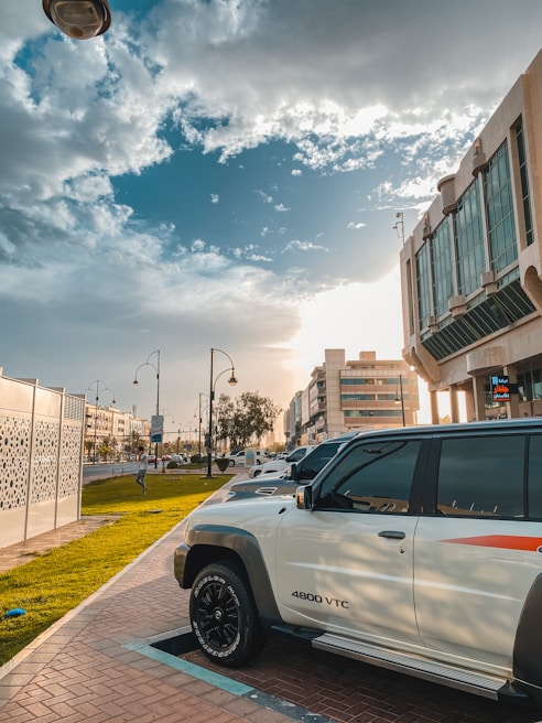 A parked white SUV with the model designation 4800 VTC is in the foreground, positioned on a paved walkway beside a grassy area. The scene is urban, featuring modern buildings and streetlights with decorative design. The sky is partly cloudy with the sun casting warm light, suggesting either morning or late afternoon.