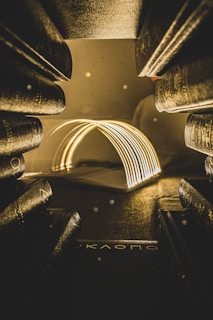An array of books is positioned around an illuminated open book, casting a warm glow in the dimly lit setting. The light emanates from the pages, creating an arch-like effect. The atmosphere is cozy and inviting, with specks of light suggesting dust particles caught in the illumination.