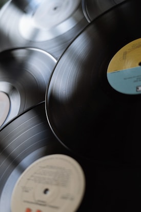 A collection of vinyl records with visible grooves and colorful labels positioned on a flat surface. The records overlap slightly, creating a layered, nostalgic look.