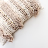 A close-up of a decorative pillow featuring intricate crochet patterns and tasselled fringes. The fabric showcases alternating rows of open stitches and densely woven textures, giving a handcrafted and artisanal appearance.