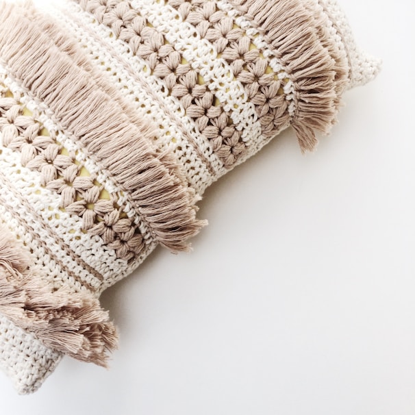 A close-up of a decorative pillow featuring intricate crochet patterns and tasselled fringes. The fabric showcases alternating rows of open stitches and densely woven textures, giving a handcrafted and artisanal appearance.
