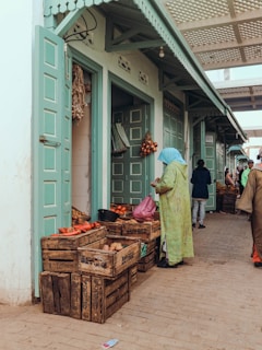 A market scene with green doors and wooden crates filled with vegetables like tomatoes and potatoes. A person wearing a blue headscarf and green robe is standing near the crates. The setting appears to be outdoors with an overhang providing shade.