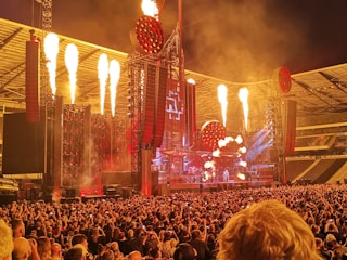 A large concert stadium with a massive crowd gathered in front of a stage. The stage setup includes elaborate light structures and pyrotechnics with flames shooting upward. The atmosphere is energetic with bright lights and red accents.