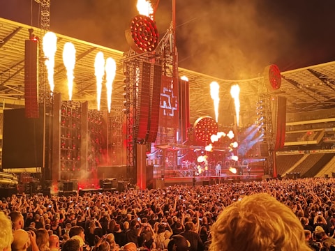 A large concert stadium with a massive crowd gathered in front of a stage. The stage setup includes elaborate light structures and pyrotechnics with flames shooting upward. The atmosphere is energetic with bright lights and red accents.