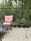 A patio area featuring a metal chair with a pink cushion, accompanied by a matching table with a glass top. Several potted plants line the border, set against a lattice fence adorned with green climbing vines.