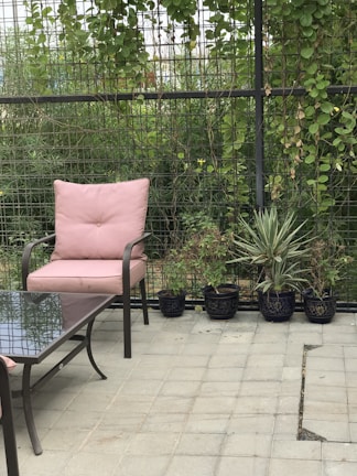 A patio area featuring a metal chair with a pink cushion, accompanied by a matching table with a glass top. Several potted plants line the border, set against a lattice fence adorned with green climbing vines.