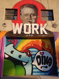 A vibrant street mural features a stylized portrait of a man within a large red circle at the top, with the word 'WORK' in bold white letters below. Below the portrait, a colorful cartoon character with a speech bubble saying 'DIVA' is depicted. The mural is painted on a roll-up door, combining abstract shapes and bright colors.