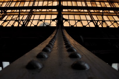 A prominent metal beam, adorned with a line of evenly spaced rivets, extends upward towards a brightly lit industrial ceiling. The overhead structure consists of large, rectangular translucent panels framed by a grid of dark metal supports, which contrast with the warm glow filtering through.