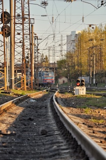 A freight train is situated on railway tracks surrounded by a complex network of overhead wires and numerous signal poles. Greenery and buildings are visible in the background, under a clear sky illuminated by warm sunlight.
