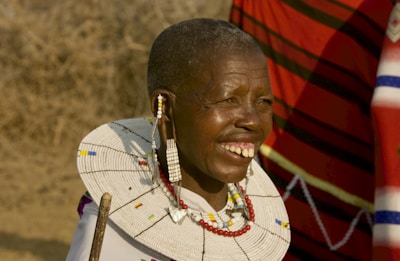 An older adult wearing a traditional Maasai garment and colorful beaded jewelry, smiling broadly. The background is a dry, textured landscape, and a red and black striped fabric is visible behind the person.
