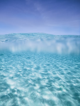 The image captures a serene view of clear, shallow ocean water. The surface creates gentle ripples, while the bottom features sandy textures visible beneath the crystal-clear water. Above the waterline, the sky meets the ocean, creating a seamless blend of blues.