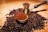A cezve filled with whole coffee beans is surrounded by scattered coffee beans on a wooden surface. In front of it, there is a bowl containing ground coffee, adding a rich texture to the scene. The lighting highlights the glossy, dark brown color of the beans and the metal shine of the cezve.