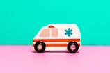A toy ambulance with a white body and red stripes is positioned against a bright turquoise backdrop. The toy features black wheels and an emergency medical symbol on the side, sitting on a vivid pink surface.