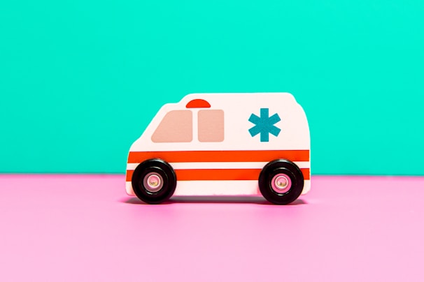 A toy ambulance with a white body and red stripes is positioned against a bright turquoise backdrop. The toy features black wheels and an emergency medical symbol on the side, sitting on a vivid pink surface.