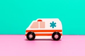 A toy ambulance with a white body and red stripes is positioned against a bright turquoise backdrop. The toy features black wheels and an emergency medical symbol on the side, sitting on a vivid pink surface.