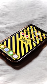 A smartphone with a colorful case displaying a patterned wallpaper featuring diagonal yellow and black stripes. The home screen shows several app icons including YouTube, Settings, and a music app.