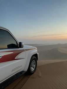 A white vehicle with red detailing is parked on a sand dune, while tracks are visible leading to and from its tires. The desert landscape stretches into the distance, with soft ripples in the sand under a pastel-colored sky, suggesting either sunrise or sunset.