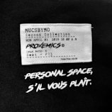 A fabric label features text mimicking a movie ticket with details such as a fictional collection name, date, time, movie title 'PROXEMICS', and seat information. Below the label, bold white text on a black background reads 'PERSONAL SPACE, S'IL VOUS PLAÎT.'