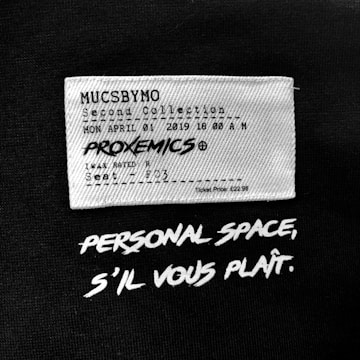 A fabric label features text mimicking a movie ticket with details such as a fictional collection name, date, time, movie title 'PROXEMICS', and seat information. Below the label, bold white text on a black background reads 'PERSONAL SPACE, S'IL VOUS PLAÎT.'