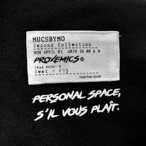 A fabric label features text mimicking a movie ticket with details such as a fictional collection name, date, time, movie title 'PROXEMICS', and seat information. Below the label, bold white text on a black background reads 'PERSONAL SPACE, S'IL VOUS PLAÎT.'