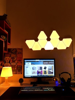 A well-organized computer desk setup features a monitor displaying various images, flanked by a pair of small speakers and a headset. The desk is illuminated by a warm glowing lamp on the left. Above the monitor, a hexagonal-shaped wall light creates a unique pattern, adding to the cozy atmosphere. Posters are visible on the wall, enhancing the personal touch of the workspace.