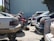 A row of cars is parked closely together in a lot, with some appearing to be missing tires, indicating a scrapyard or salvage yard setting. The cars are positioned in front of a large, corrugated metal building. Some trunks are open, and a transmission or automotive part lies on the ground.