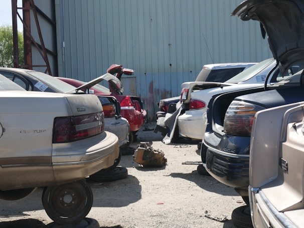 A row of cars is parked closely together in a lot, with some appearing to be missing tires, indicating a scrapyard or salvage yard setting. The cars are positioned in front of a large, corrugated metal building. Some trunks are open, and a transmission or automotive part lies on the ground.