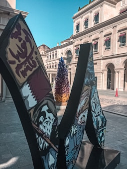An outdoor scene featuring a modern sculpture with urban art and calligraphy. The sculpture includes a depiction of a figure, possibly historical or mythical, with intricate designs. A vibrant, colorful installation resembling a tree or abstract art piece stands in the background. Classic architecture surrounds the plaza with beige stone buildings and arched windows. The sky above is clear and blue.