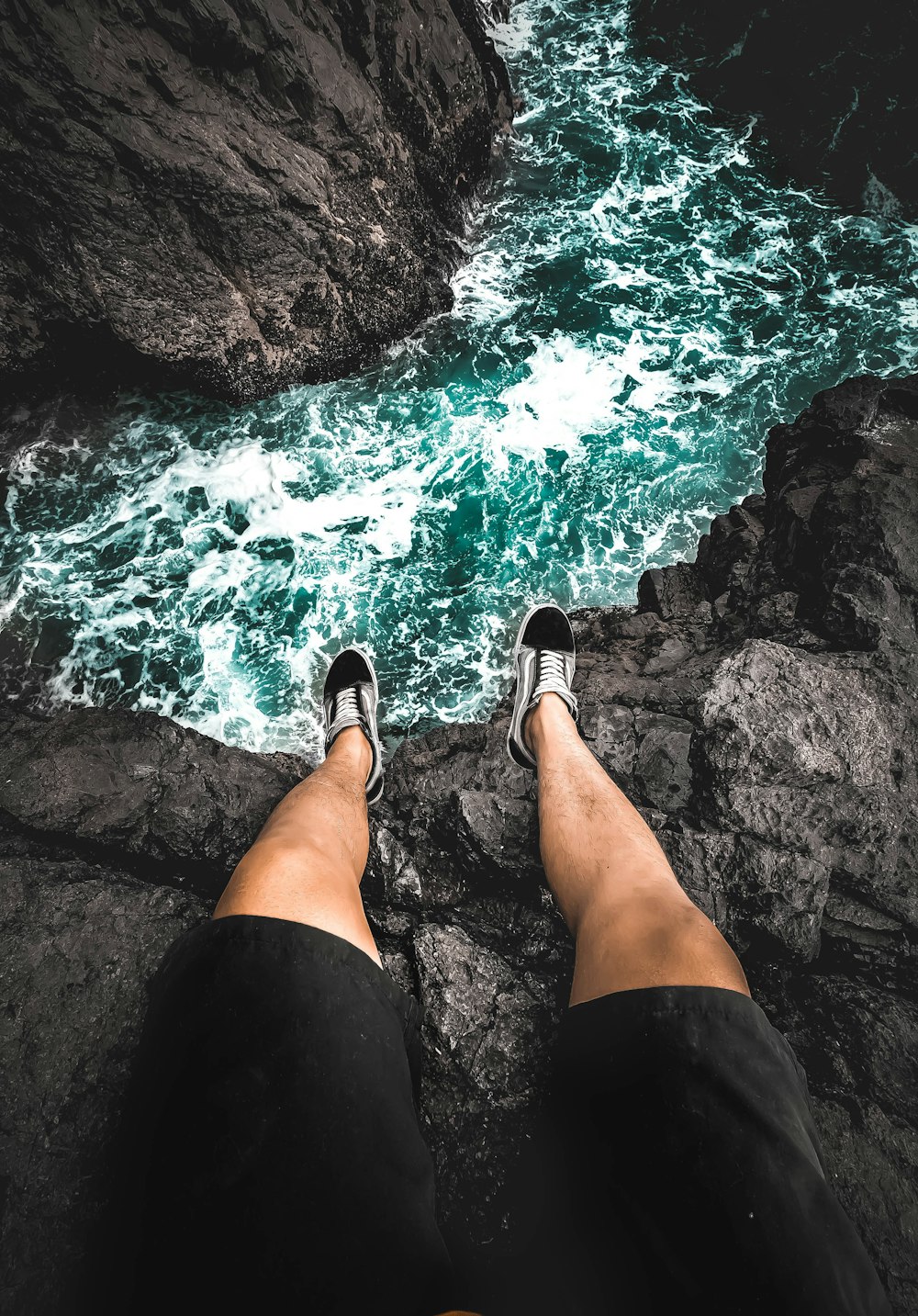 person in black shorts and black shoes sitting on rock near body of water during daytime