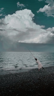 A person in a striped outfit is fishing by the shore, casting a long fishing rod into the sea. The waves gently lap at the pebbled beach, while large, dramatic clouds fill the sky above, creating a contrast with the dark water.