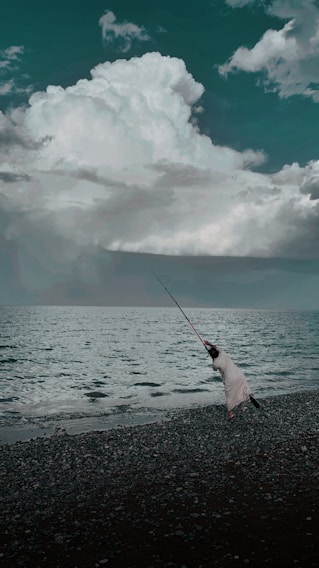 A person in a striped outfit is fishing by the shore, casting a long fishing rod into the sea. The waves gently lap at the pebbled beach, while large, dramatic clouds fill the sky above, creating a contrast with the dark water.