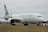A large passenger airplane with 'Air New Zealand' branding is positioned on an airport runway. The aircraft, in a white and black color scheme, is stationary and surrounded by flat terrain with some trees in the background. The sky is overcast, contributing to a neutral atmosphere.