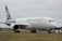A large passenger airplane with 'Air New Zealand' branding is positioned on an airport runway. The aircraft, in a white and black color scheme, is stationary and surrounded by flat terrain with some trees in the background. The sky is overcast, contributing to a neutral atmosphere.