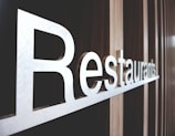 Large metallic letters spelling out 'Restaurants' are mounted on a textured wooden wall. The perspective of the photo highlights the length and sleekness of the letters, creating a modern and inviting atmosphere.