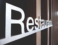 Large metallic letters spelling out 'Restaurants' are mounted on a textured wooden wall. The perspective of the photo highlights the length and sleekness of the letters, creating a modern and inviting atmosphere.