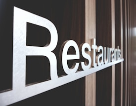 Large metallic letters spelling out 'Restaurants' are mounted on a textured wooden wall. The perspective of the photo highlights the length and sleekness of the letters, creating a modern and inviting atmosphere.
