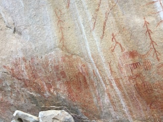 Ancient rock art featuring a series of red ochre figures and patterns painted on a large rock surface. The figures appear to depict human-like forms and various geometric shapes. The artwork suggests a historical or cultural significance, potentially related to human activity or rituals.