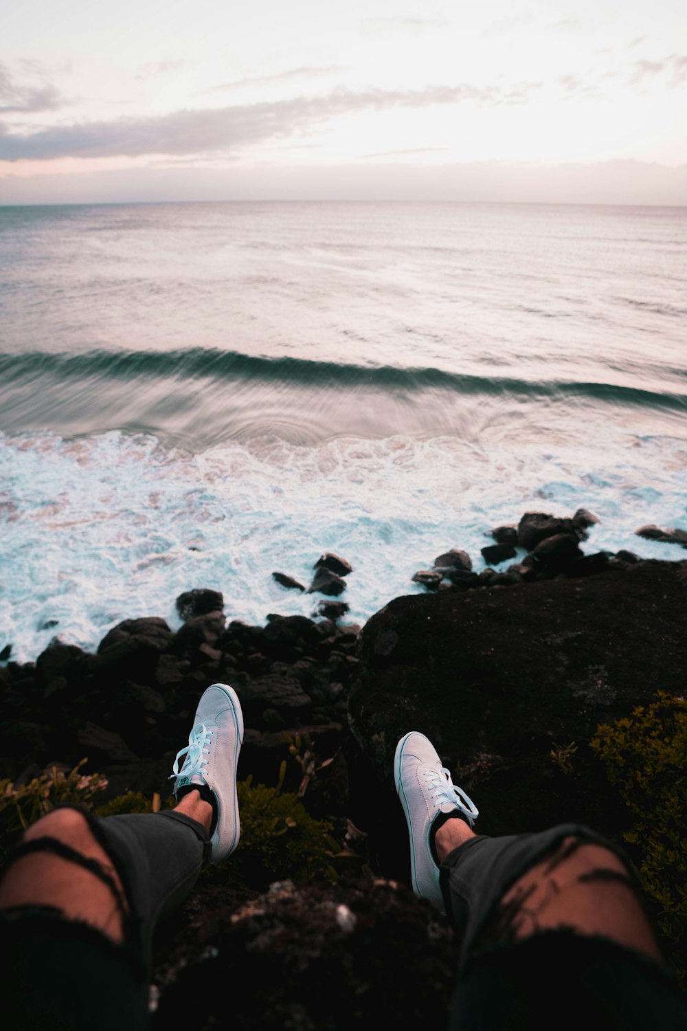 person in black shorts and white sneakers sitting on rock by the sea during daytime